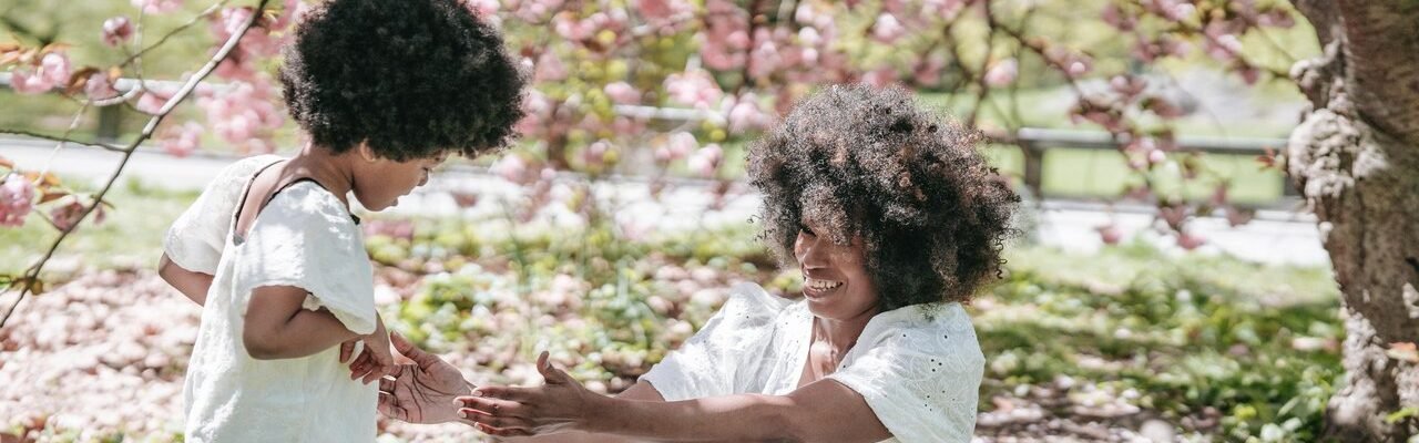 Madre e hija con vestido blanco y cabello chino. En un campo, bajo la sombra de un árbol.